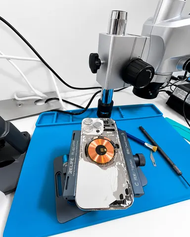 An iPhone with its back glass off, clamped under the stereo microscope on the Jimmy repair bench